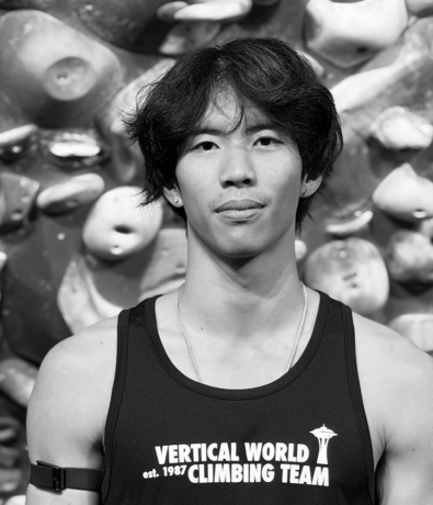 A young man wearing a Vertical World Climbing Team tank top poses in front of an indoor rock climbing wall during a bouldering project.