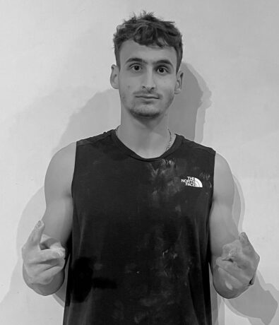 A young man in a black tank top with chalk-covered hands pointing forward during a bouldering project.