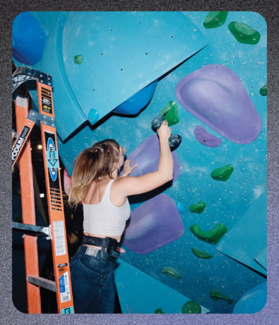 A person on a ladder uses a cordless drill to attach a green climbing hold to a blue indoor rock climbing wall for a marketing strategy.