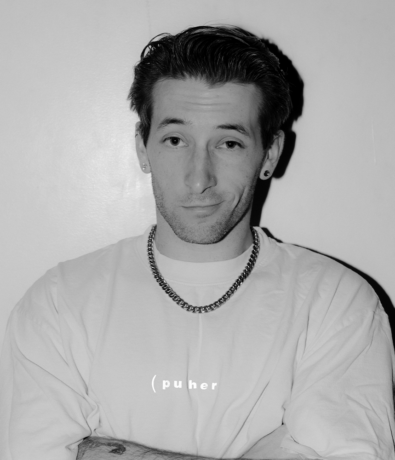 A black-and-white portrait of a man wearing a chain necklace and a t-shirt with puher printed on the chest, captured during a Bouldering Project community climbing competition.