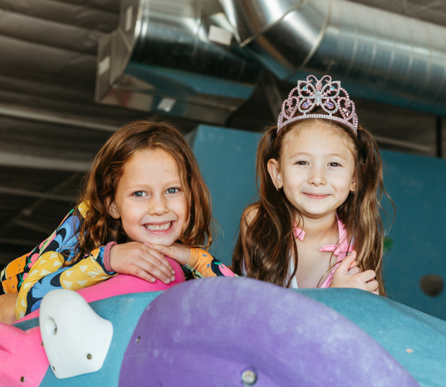 Two young girls smile while leaning on a climbing wall during Austin youth programs, one wearing a pink tiara.