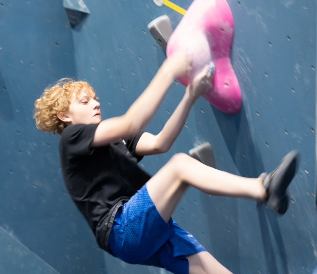 A young climber at a Tempe youth climbing club reaches for a large pink hold on a blue indoor wall.