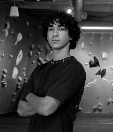 A person with curly hair stands with their arms crossed in front of a climbing wall during a community bouldering project.