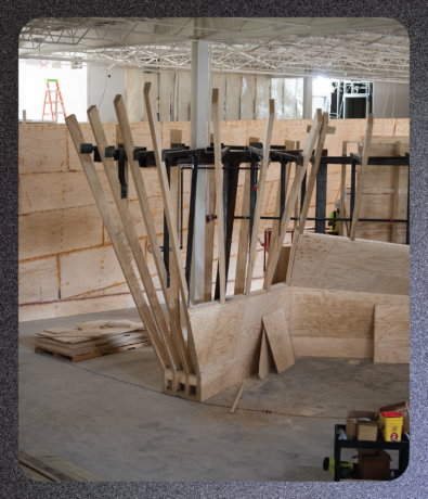 A construction site featuring a framework of vertical plywood boards surrounding a central metal pillar, with plywood wall panels and a ladder in the background, serving as a visual for a brand campaign.