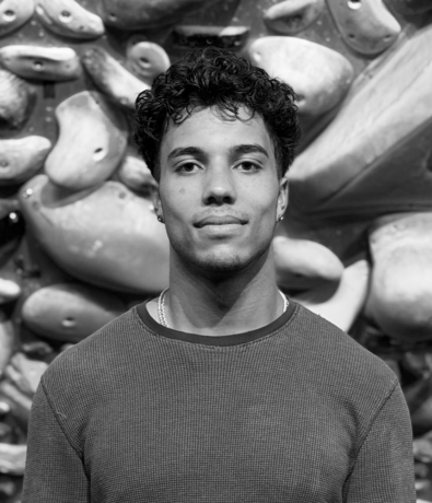 Portrait of a young man in a long-sleeved shirt standing in front of a rock climbing wall at a bouldering project.