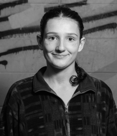 A black-and-white portrait of a smiling person in a zip-up fleece jacket against a graffiti-covered wall at the Bouldering Project.