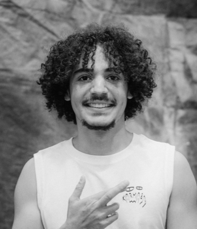 A young person with curly hair smiling and pointing toward their chest at the Bouldering Project.