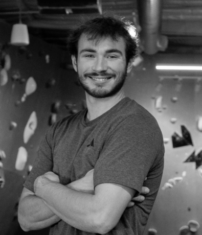 A young man with a beard stands with his arms crossed in front of a climbing wall at the bouldering project.