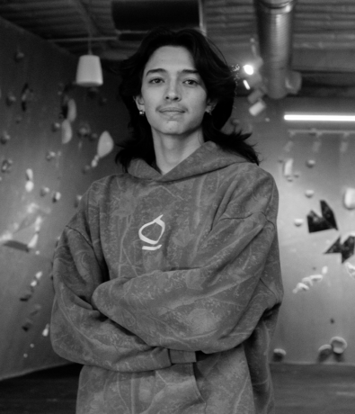 A person with long dark hair stands with arms crossed at a bouldering project in a climbing gym.