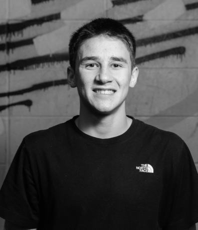 A smiling young man in a black T-shirt stands in front of a graffiti-covered brick wall at a bouldering project community event.