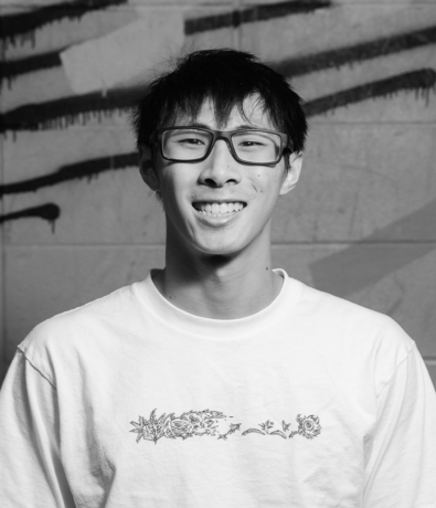 A smiling young man in a white graphic t-shirt and glasses stands in front of a graffiti-covered wall at the Bouldering Project.