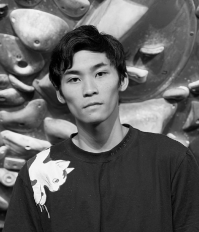 A young man in a black t-shirt standing in front of an indoor bouldering project wall.