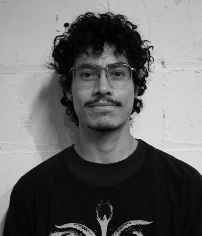 Portrait of a young person with curly hair and glasses, wearing a sweatshirt, posing in front of a white brick wall at a Bouldering Project climbing competition.
