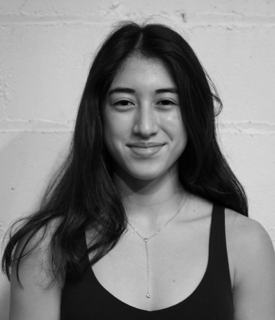 A young woman with long dark hair smiles at the camera while at a bouldering project, wearing a necklace with a small pendant.