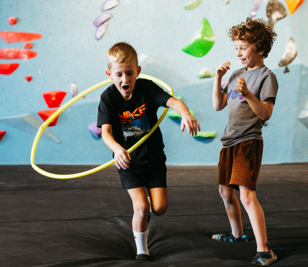 Two boys play with a yellow hula hoop in front of an indoor rock climbing wall at one of our youth climbing clubs.