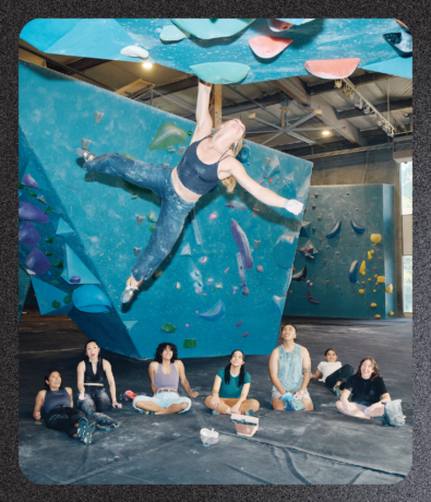 A woman suspended mid-air on a blue climbing wall as six people sit on the mat below, watching her as part of this brand campaign.