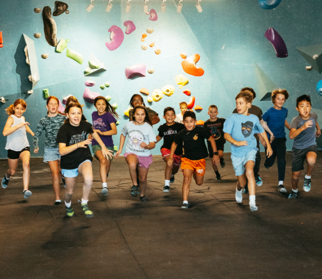 A group of children participating in Austin youth programs run together across the floor of an indoor rock climbing gym.