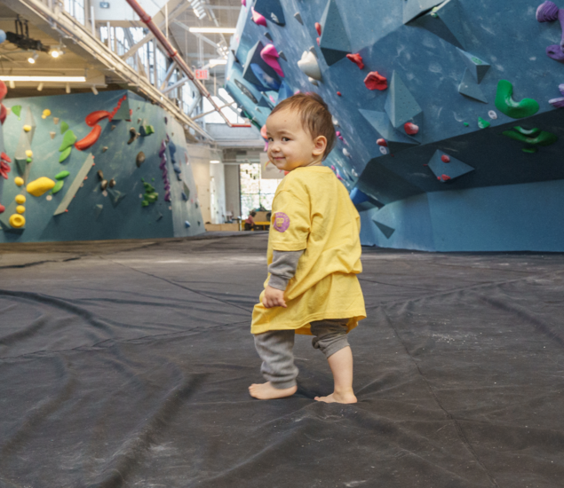 A toddler in a yellow shirt walks barefoot on the soft floor of a Tempe climbing gym.