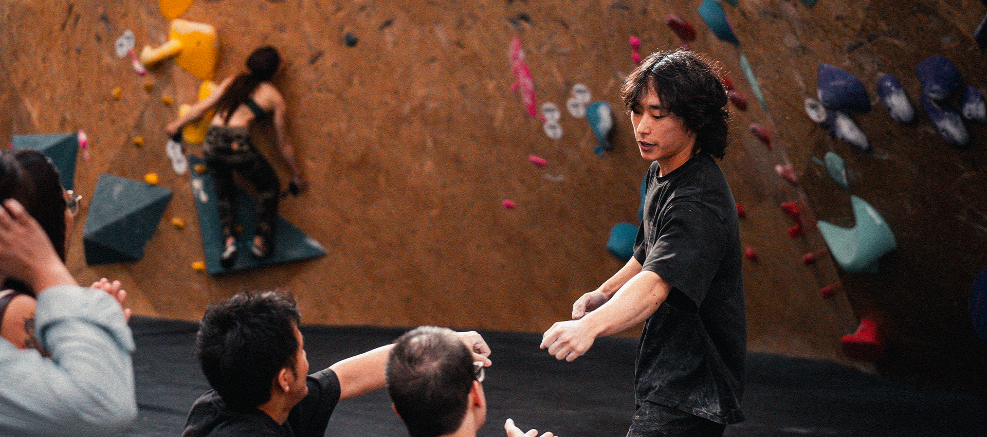 A group of people at a Boston indoor climbing gym observe a climber on a wall with colorful holds.