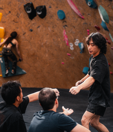 A man gestures while talking to two others on a Boston indoor climbing gym floor, with a person bouldering on a wall in the background.