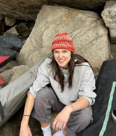 A smiling life coach with long dark hair wears a red and orange knit beanie and grey sweatshirt while sitting among large rocks and climbing crash pads in Salt Lake City.