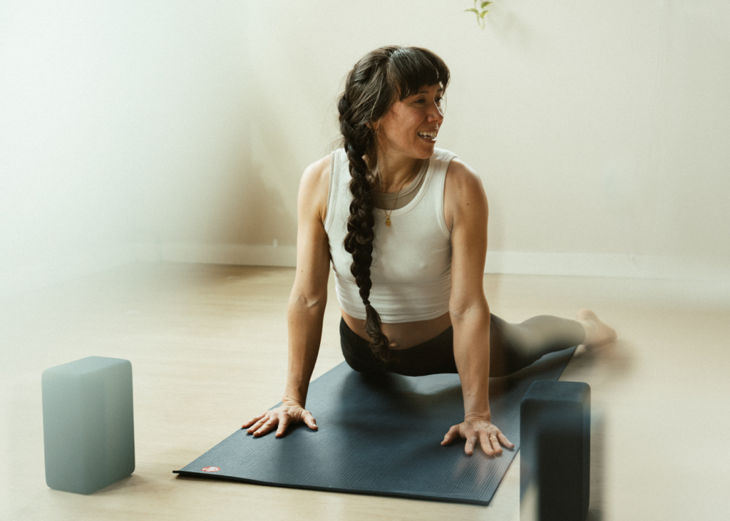 A woman with a long braided ponytail smiles while practicing cobra pose on a mat for beginners Pilates, with two yoga blocks nearby.