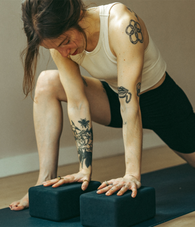A person with tattooed arms performs a low lunge yoga pose for beginners Pilates, resting their hands on two black yoga blocks.