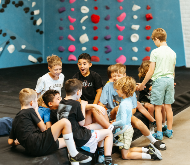 A group of children participating in Austin youth programs gathered on a floor mat in front of a climbing wall.